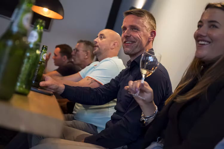 Vijf medewerkers van Haafkes zitten samen aan een houten bar, sommigen met glazen bier en wijn, lachend en genietend.