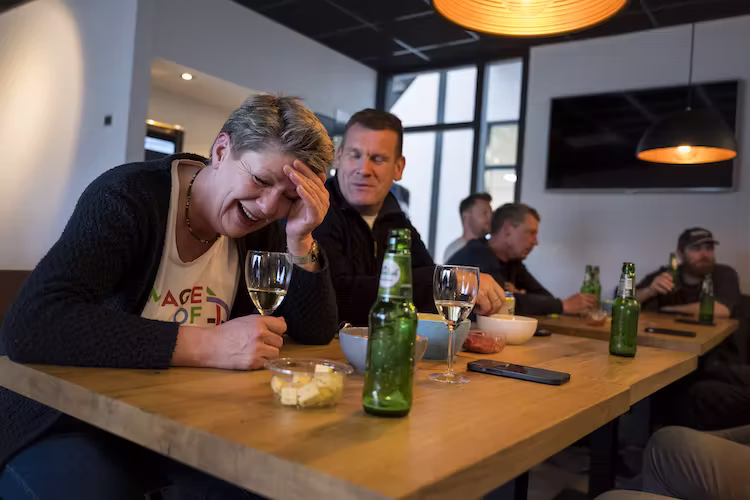Hetty van Haafkes die lacht met haar hand op haar voorhoofd terwijl ze aan een tafel zit met een glas wijn en een fles bier, omringd door andere medewerkers die ook drinken en praten in een gezellige binnenruimte.