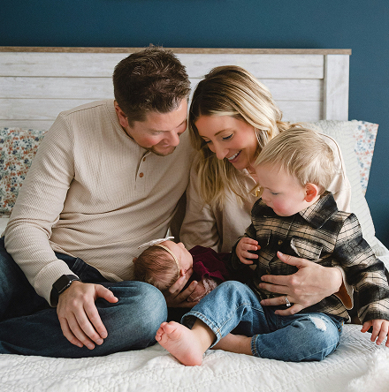 A loving family of four sits on a bed, smiling down at a newborn baby held by the mother. A toddler in plaid sits beside them, creating a warm, joyful scene.