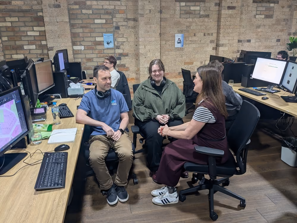 Three people seated in office chairs having a discussion in an open-plan workspace with computers.