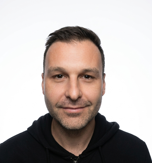 Portrait of a man, Sean, with short dark hair wearing a black polo shirt, smiling slightly against a white background.
