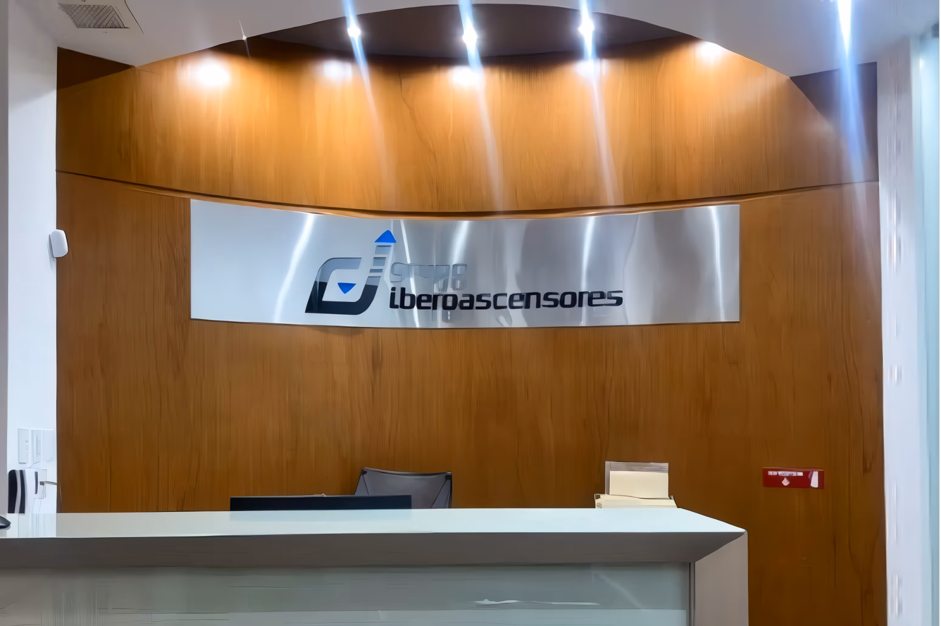 Área de recepción con un mostrador blanco y una pared curva de madera en la que se encuentra el logotipo de "GRUPO iberoascensores" en metal cepillado.