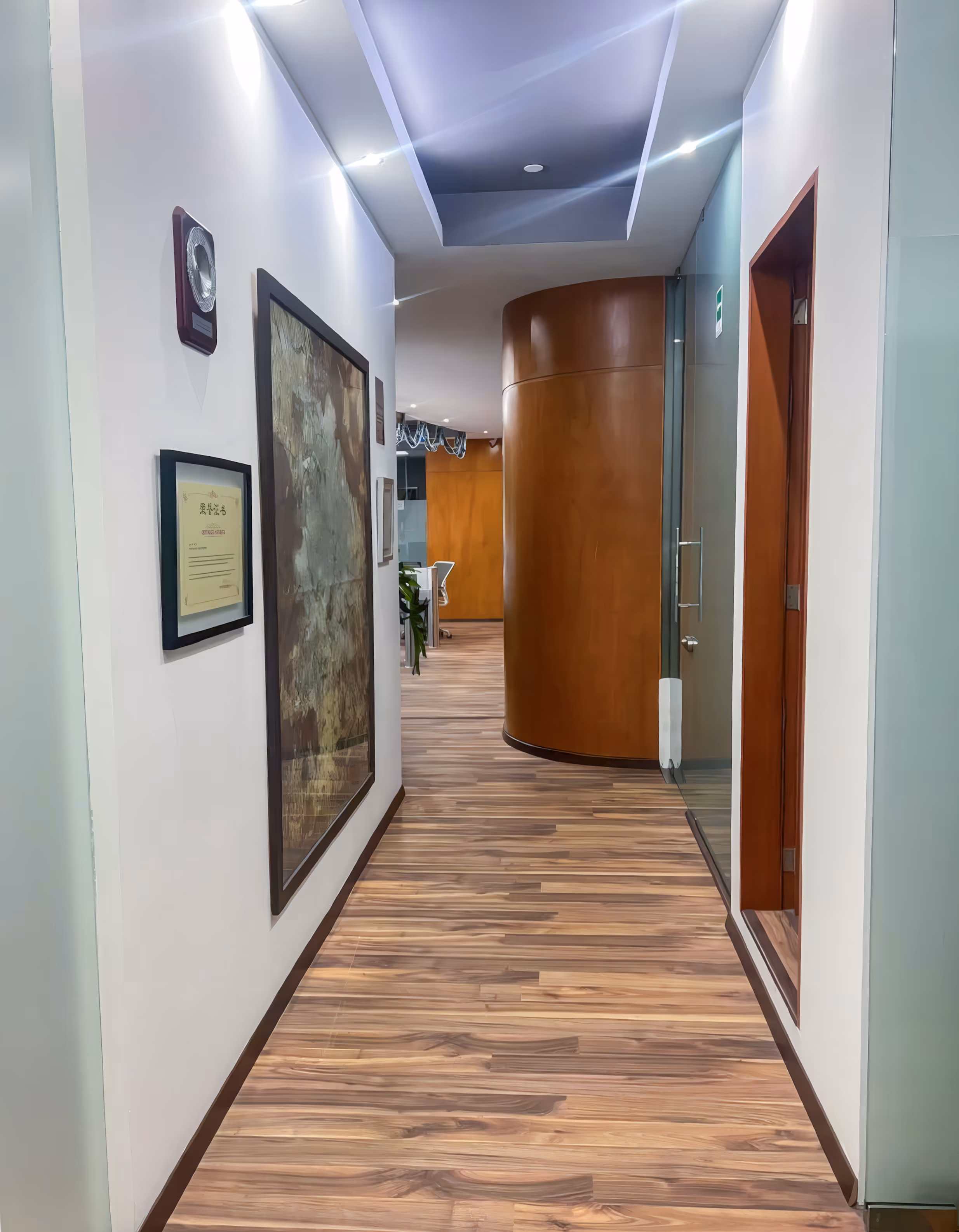 Pasillo largo de una oficina con piso de duela laminada, paredes blancas y cuadros decorativos. Al fondo se observa una pared curva de madera y el resto del área de trabajo.
