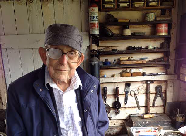An elderly gentleman stood in a workshop with a workbench and tools