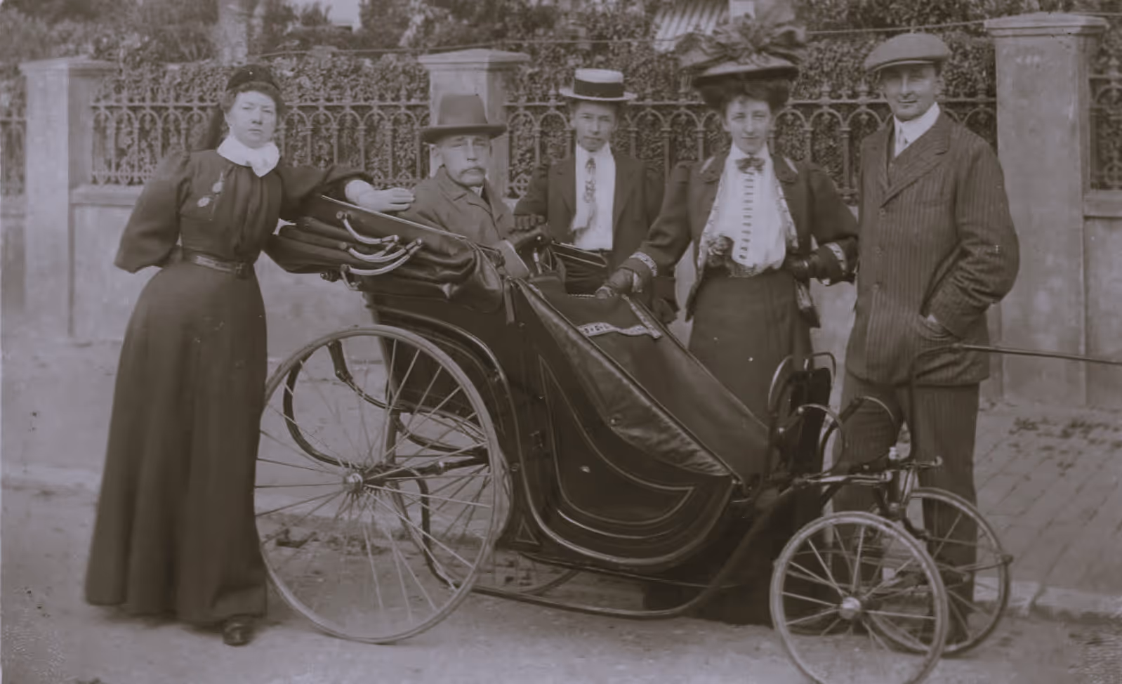 A vintage photograph of four adults standing around an early three-wheeled carriage with a seated man.