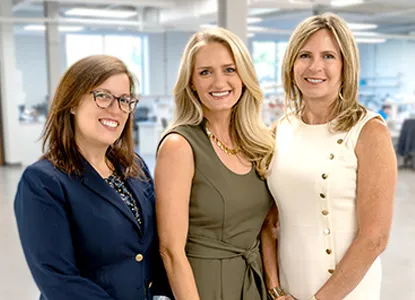 Three women smiling, standing indoors in professional attire in a well-lit office space.