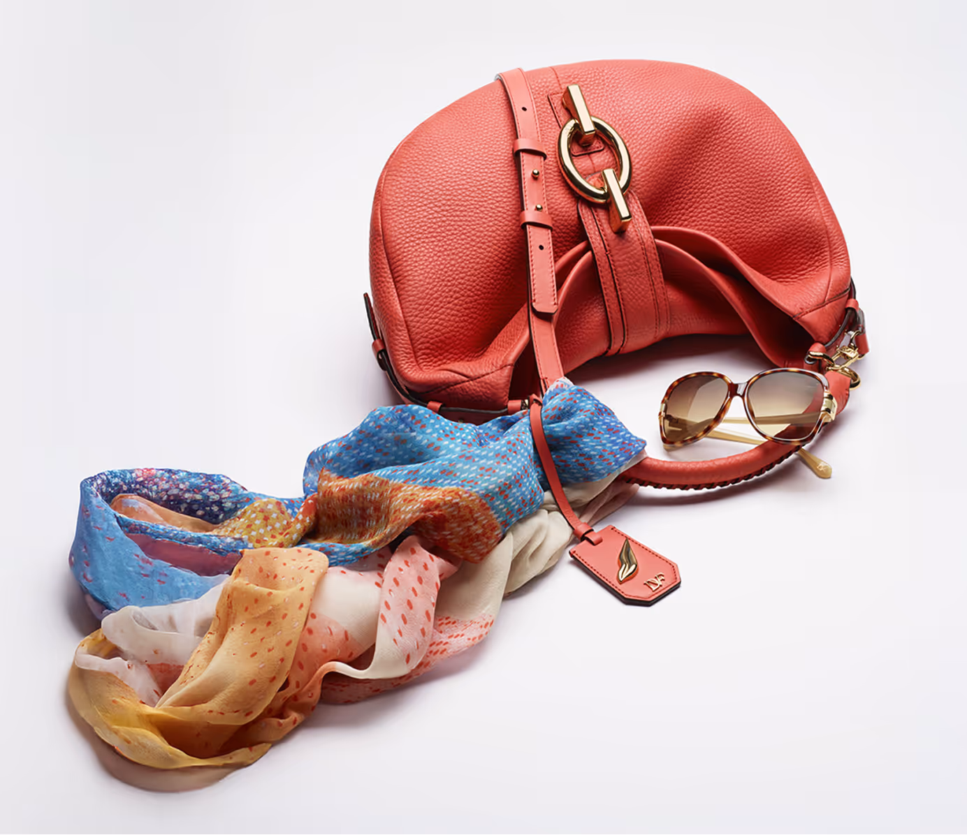 Red leather handbag with gold hardware, multicolored silk scarf, and tortoiseshell sunglasses on a white background.