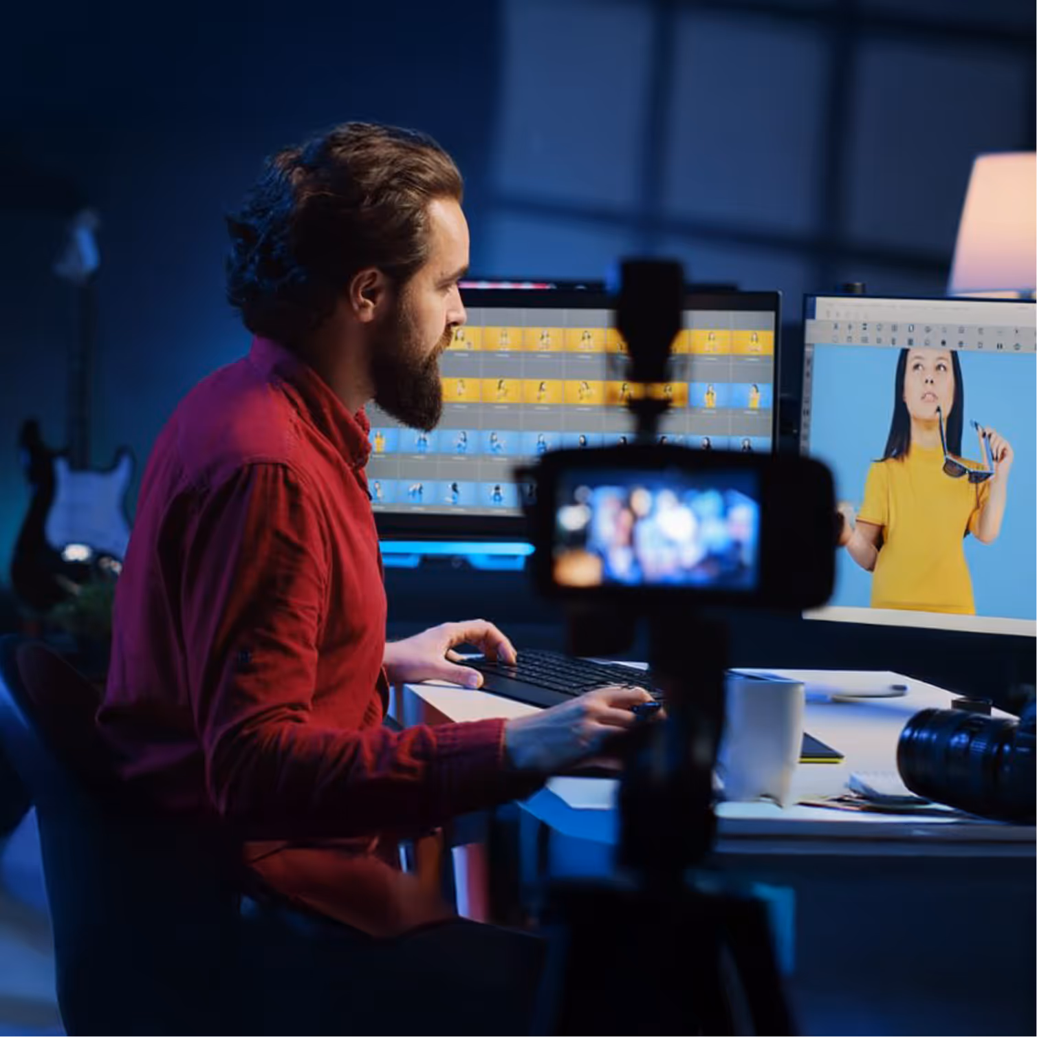 Bearded man in red shirt editing video footage on dual computer monitors in a dimly lit room with a camera recording in the foreground.