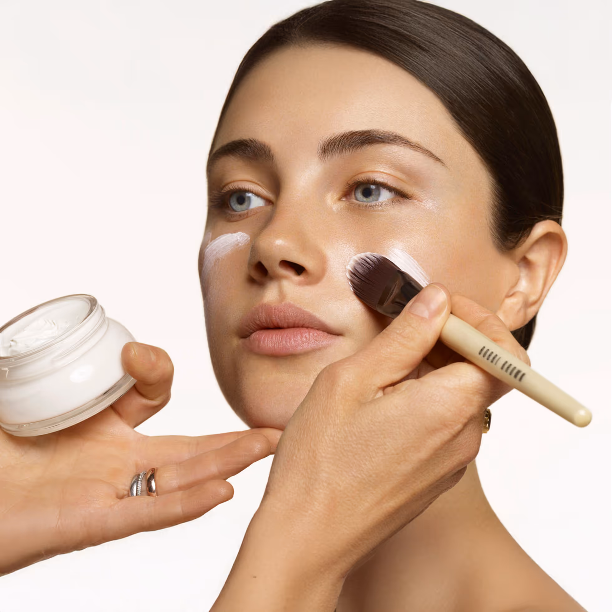 Person applying white cream on a woman's cheek with a makeup brush while holding a jar of cream.