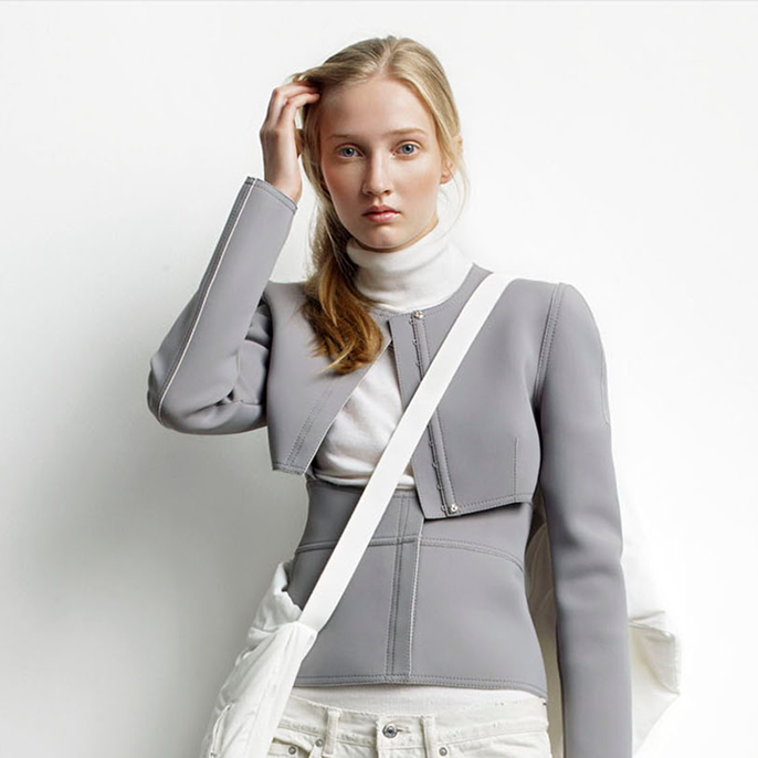 Blonde woman wearing a gray cropped jacket over a white turtleneck and carrying a white shoulder bag.