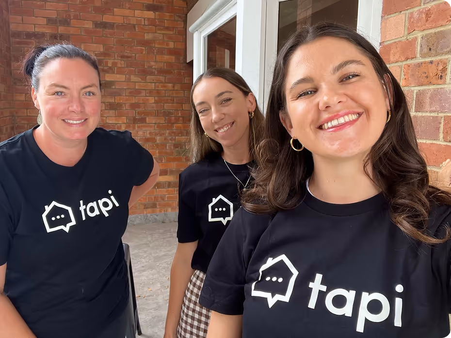 Three women wearing black Tapi t-shirts smiling together in front of a brick building.