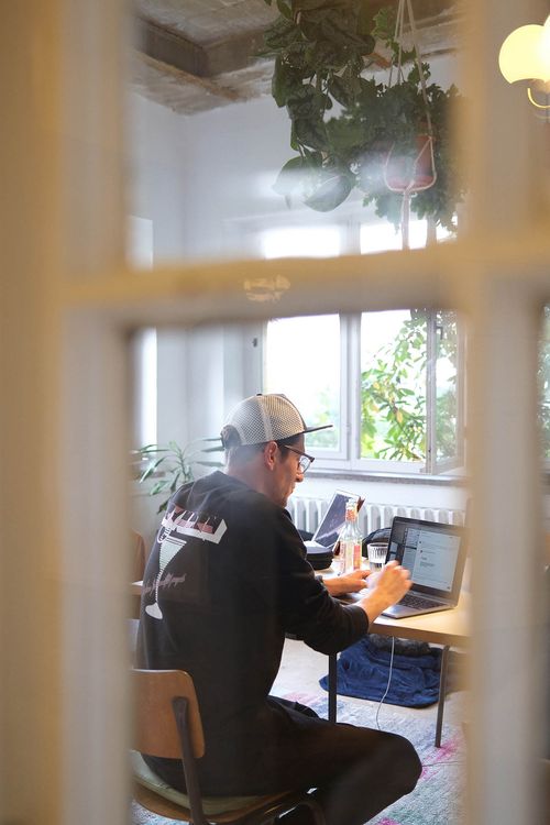 Michael Rahmfeld, Geschäftsführer von hej.build, sitzt mit einer Baseballkappe an einem Tisch in einem modernen, lichtdurchfluteten Büro, arbeitet an seinem Laptop und hat eine Getränkeflasche neben sich.