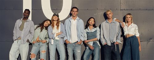 Group of seven young adults wearing casual denim and striped shirts leaning against a wall with a large 'O' painted on it.