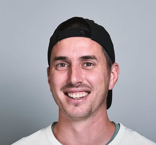 Smiling man wearing a black backward baseball cap and a light-colored shirt against a neutral gray background.