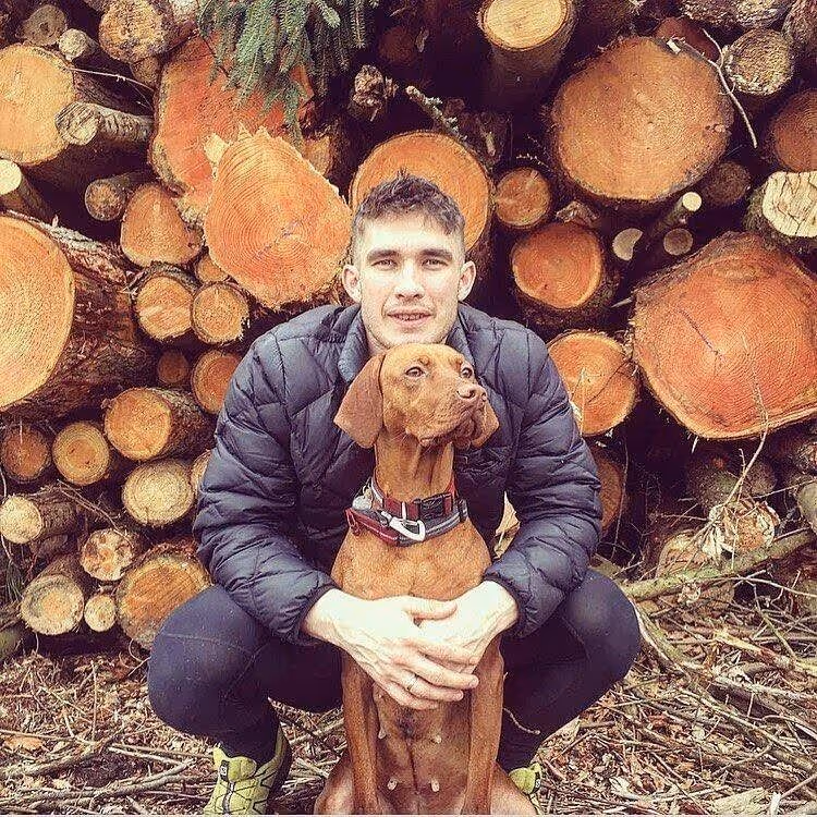 Man in a black jacket crouching and hugging a brown dog, with stacked logs in the background.