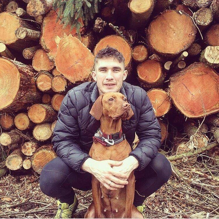 Man in a black jacket crouching and hugging a brown dog, with stacked logs in the background.