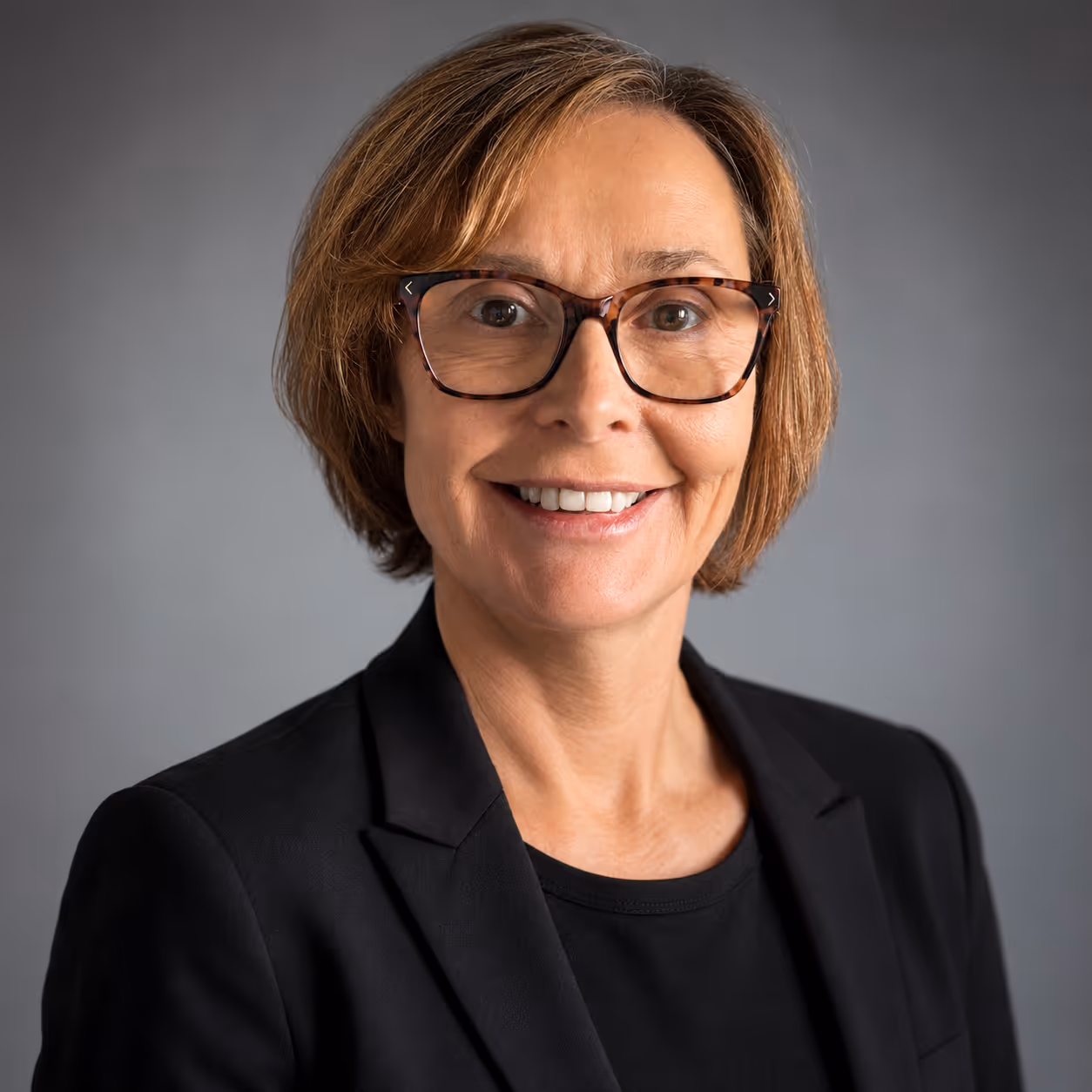 Smiling woman with short hair, wearing glasses and a black blazer against a grey background.