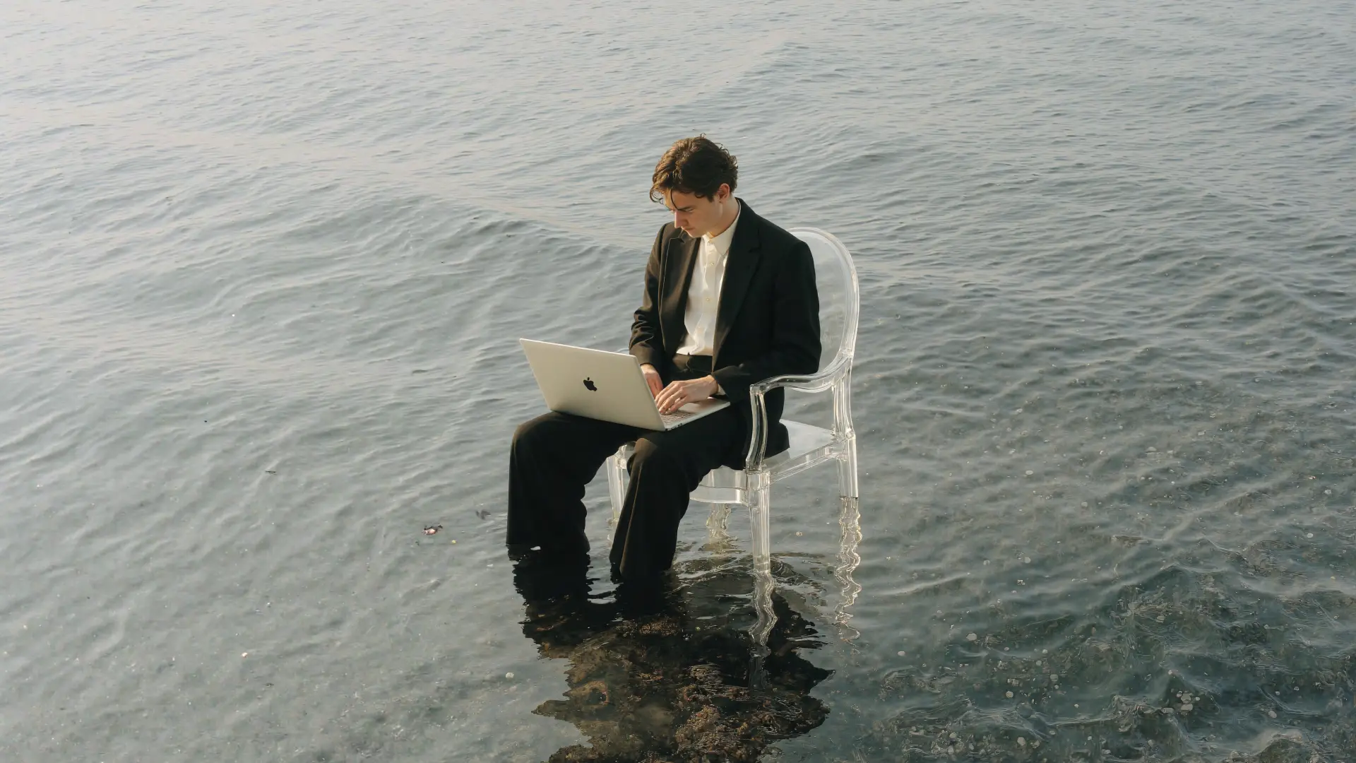 Ein Mann in einem schwarzen Anzug sitzt auf einem transparenten Stuhl im flachen Wasser und arbeitet an einem Laptop.
