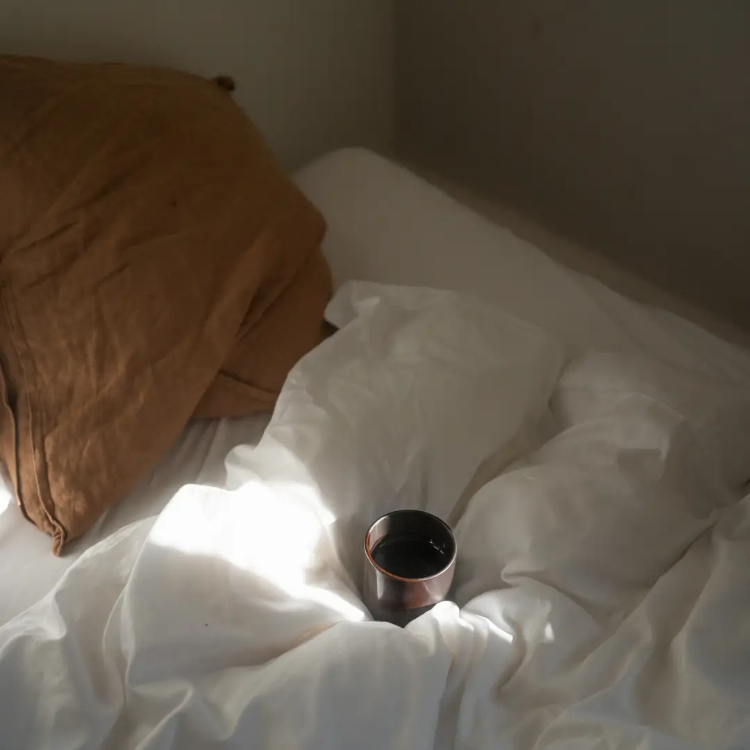 Becher mit Kaffee auf zerknitterter weißer Bettdecke neben braunem Kissen im Sonnenlicht.