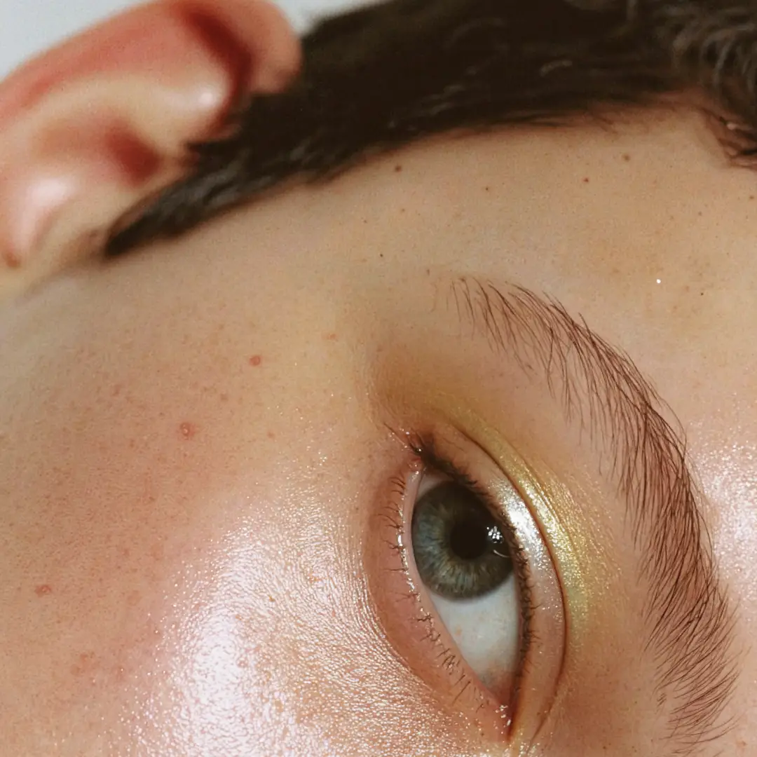 Nahaufnahme eines teilweise geöffneten grünen Auges mit goldfarbenem Lidschatten und Sommersprossen auf der Haut.