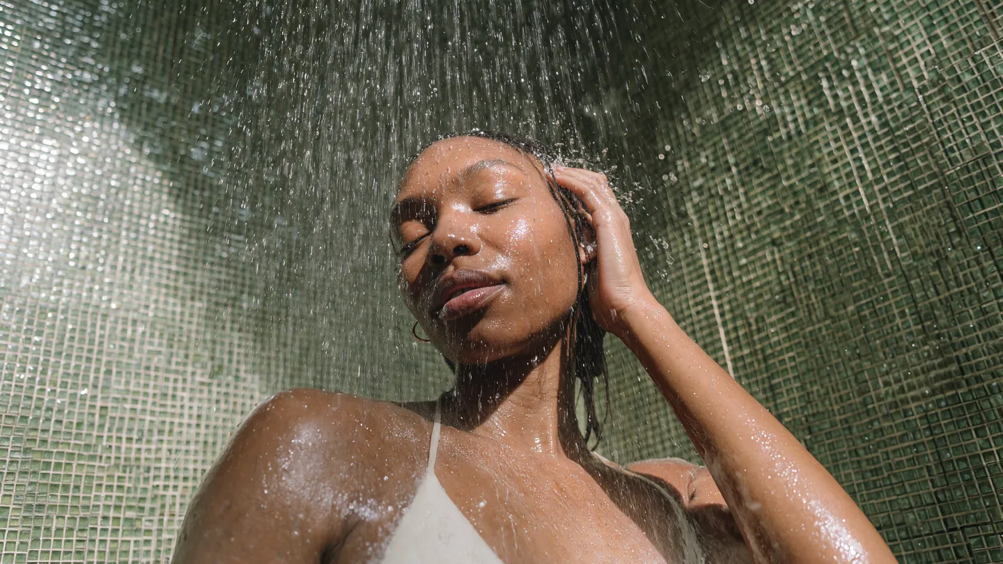 Frau mit geschlossenen Augen, die sich unter einer Dusche entspannt, während Wasser auf sie fällt.
