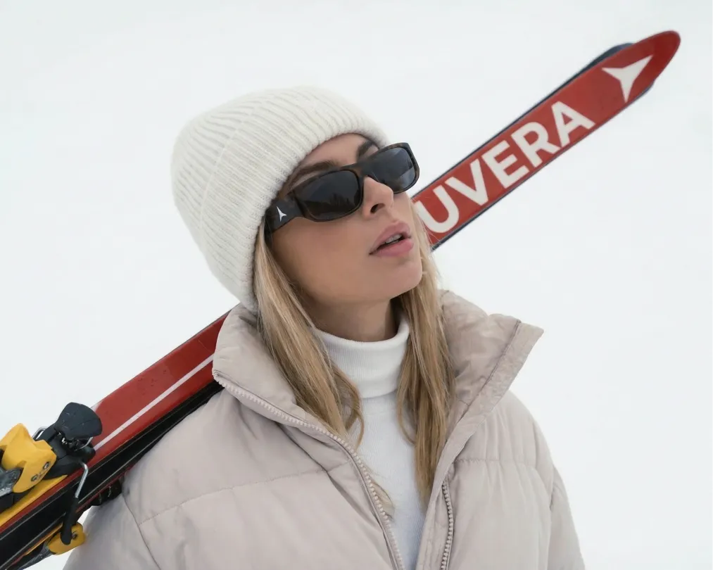 Frau mit weißer Mütze, Sonnenbrille und beige Winterjacke trägt rote Skier mit der Aufschrift UVERA auf der Schulter vor weißem Hintergrund.