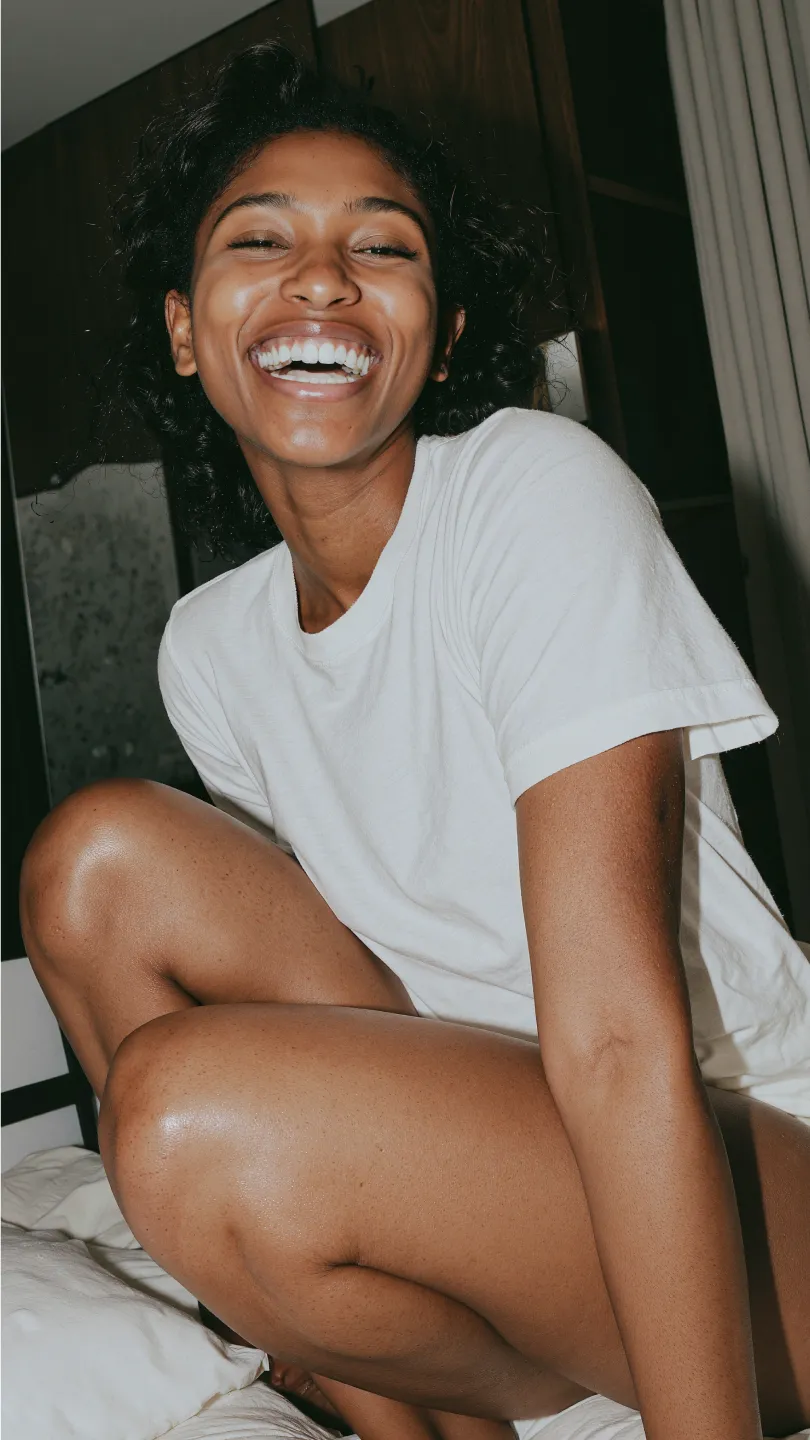 Lächende Frau mit lockigem Haar und weißem T-Shirt sitzt auf einem Bett.