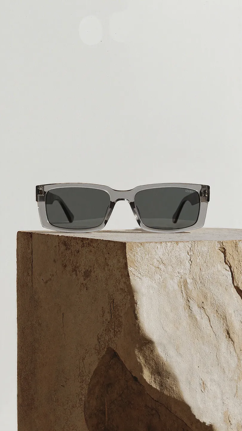 Rechteckige Sonnenbrille mit transparentem Rahmen und dunklen Gläsern auf einem groben, rechteckigen Steinstück.