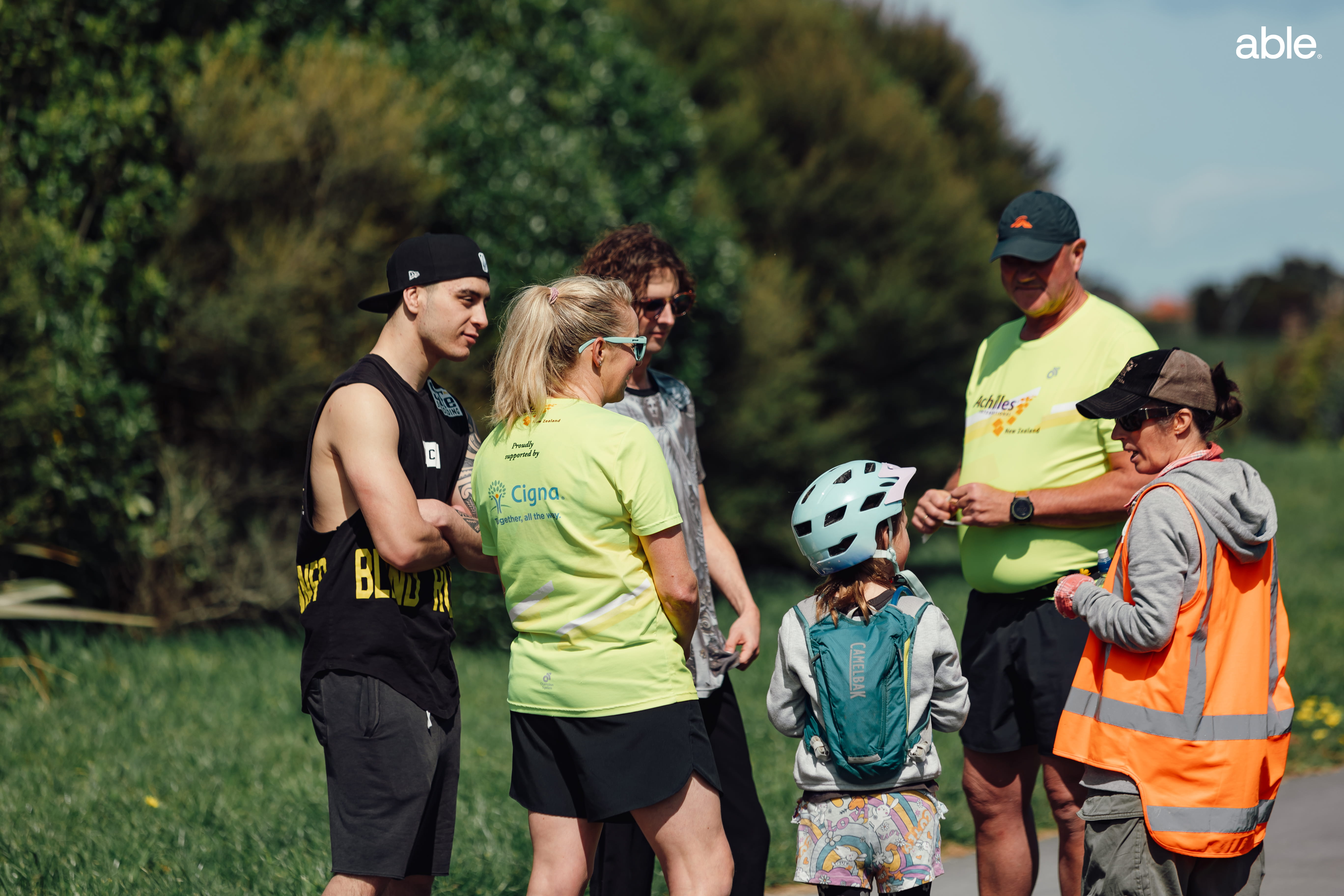 Cast, crew and volunteers stand talking in a group during filming at an outdoor running track.