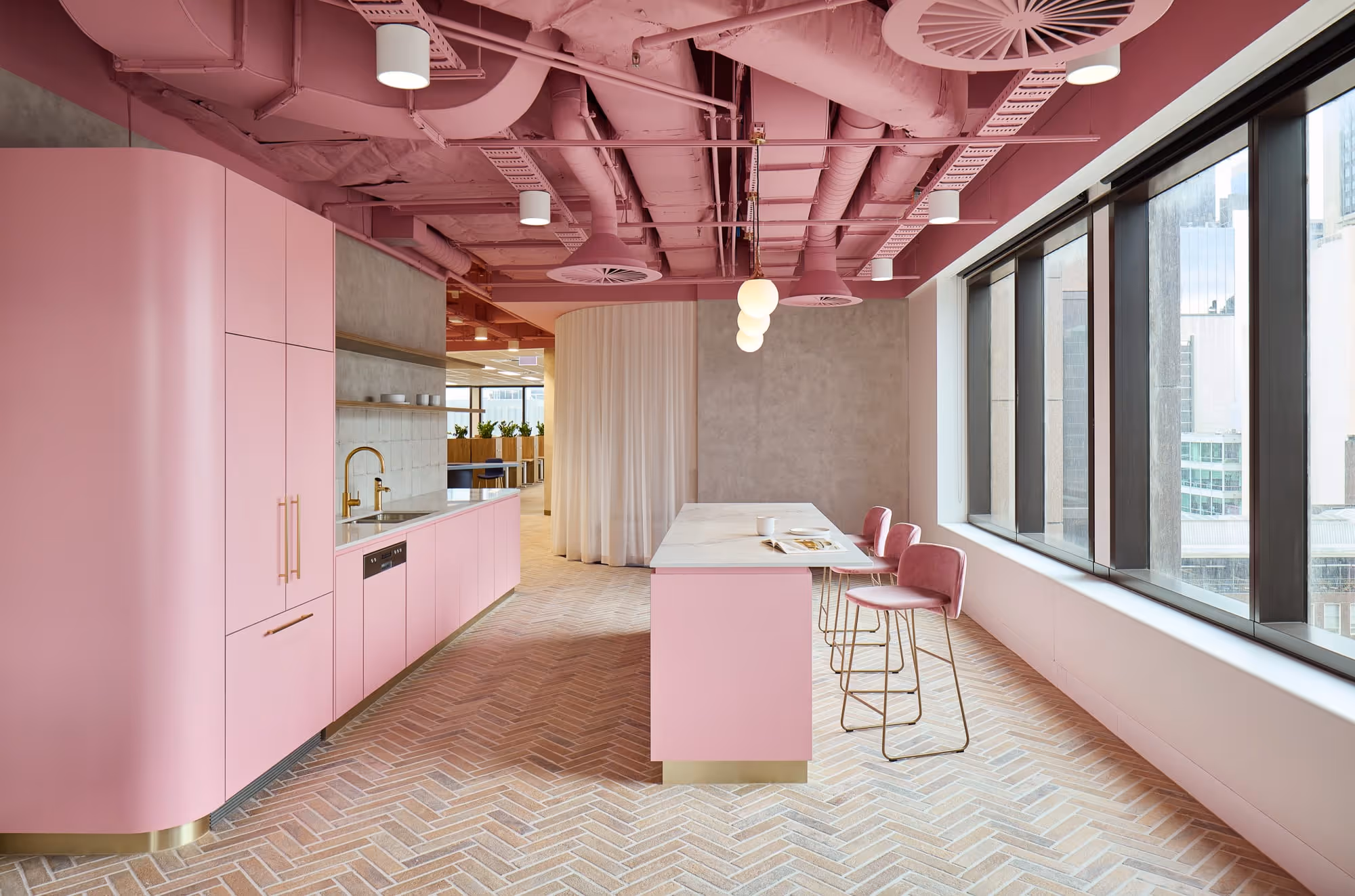Modern pink kitchen with island, velvet chairs, gold fixtures, and large windows overlooking city buildings.