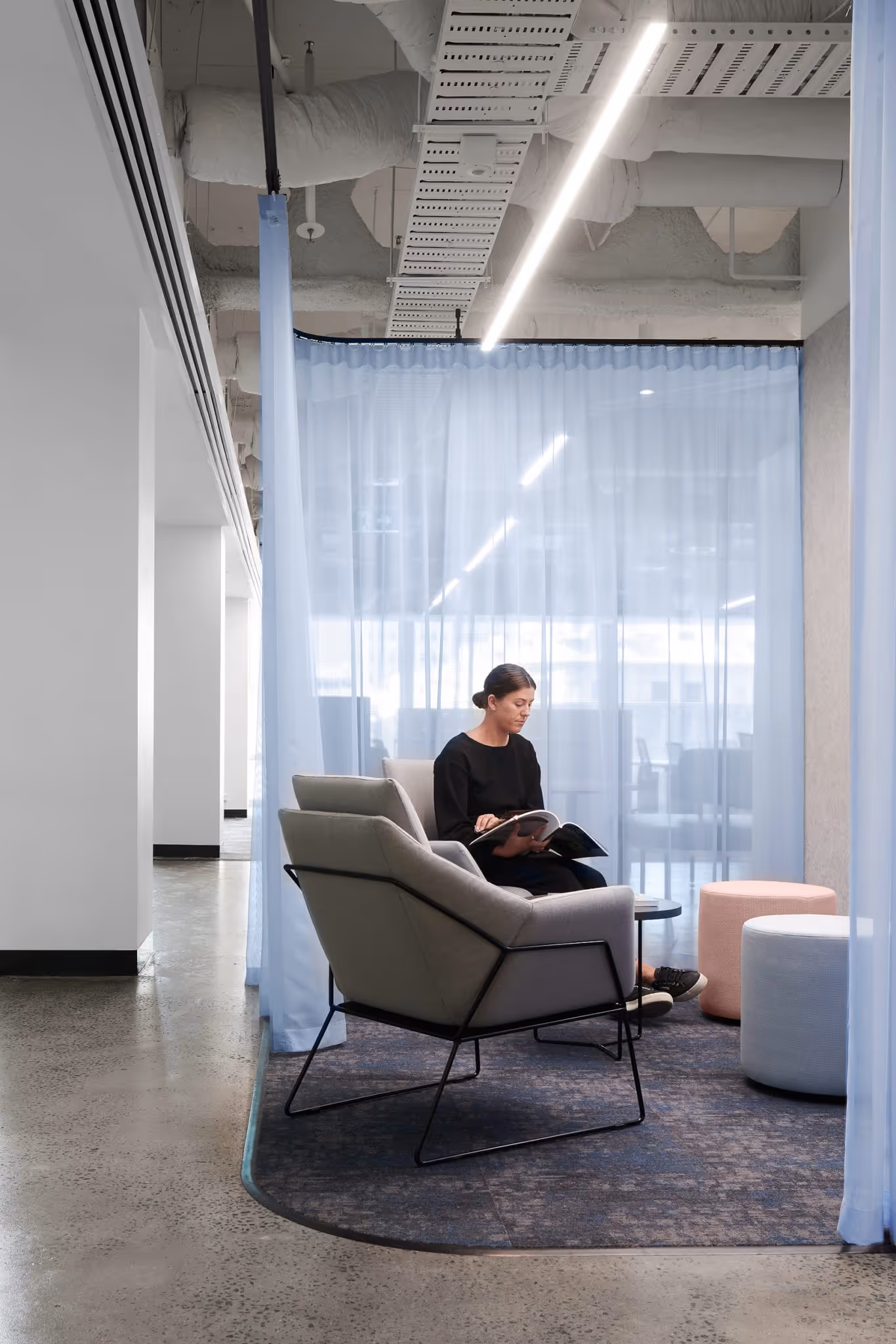 Woman in black outfit sitting on gray chair reading a magazine in a modern office lounge.
