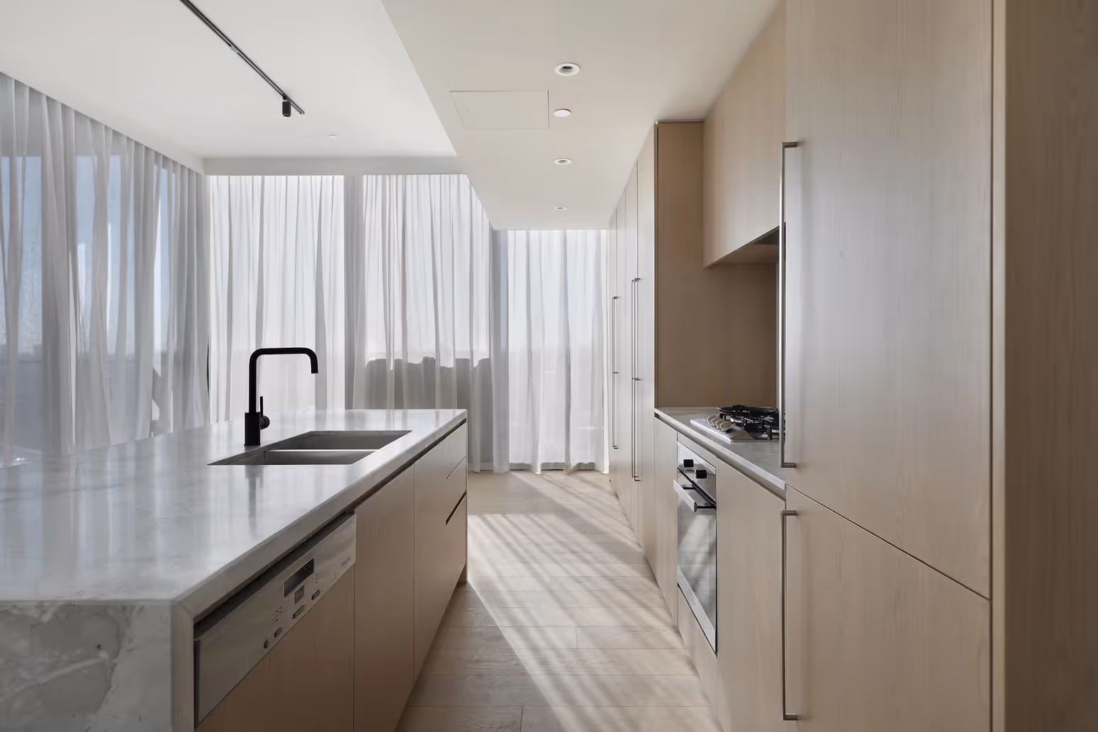Bright modern kitchen with marble island, black faucet, wood cabinets, oven, and sheer white curtains.