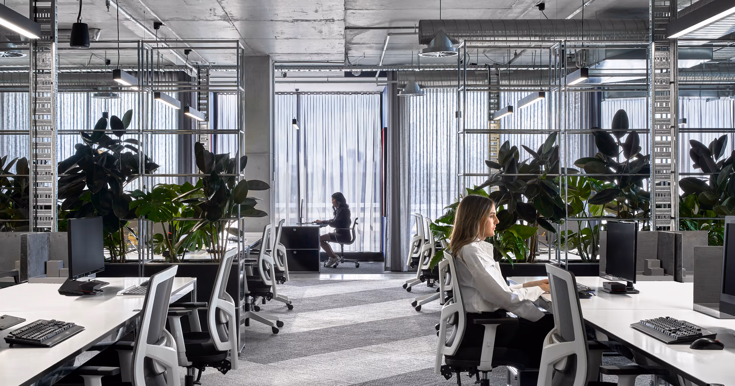 Modern office with white desks, ergonomic chairs, plants, and two women working on computers.