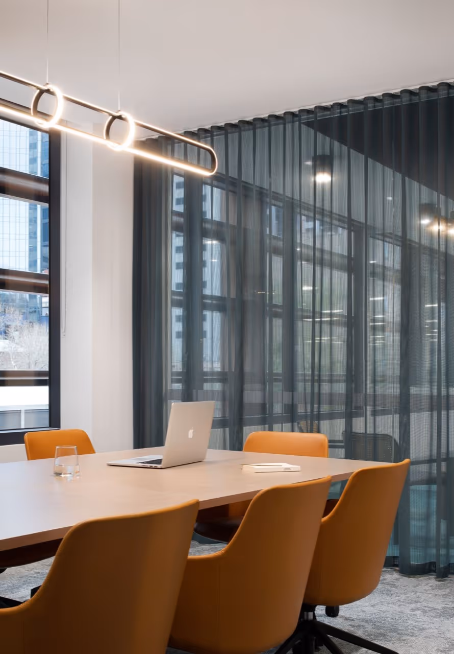 Modern office meeting room with wooden table, orange chairs, laptop, glass, and dark sheer curtains.