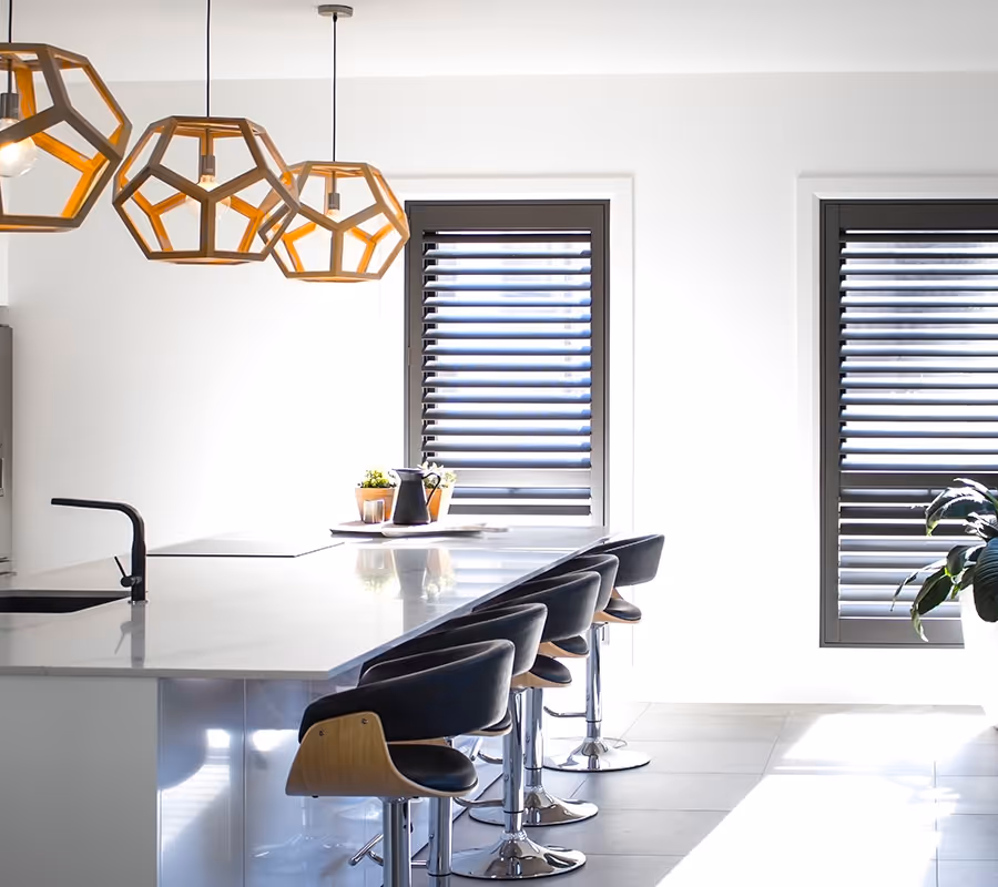 Modern kitchen island with four black bar stools, pendant lights, and windows with horizontal shutters.