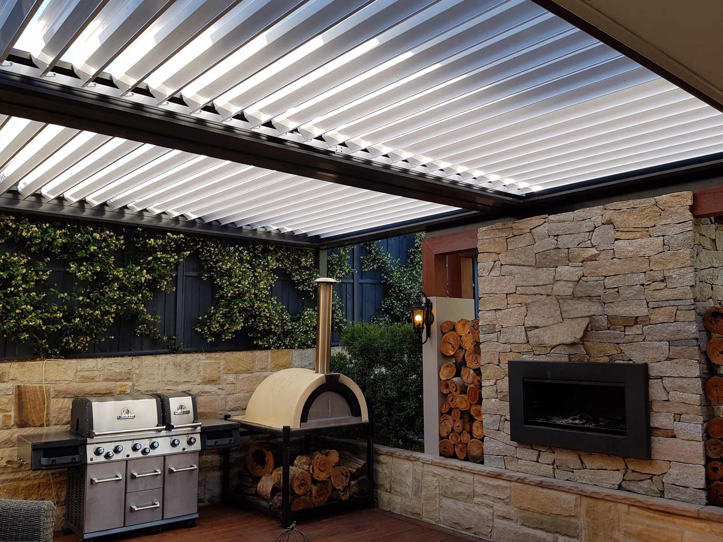 Outdoor kitchen with stainless steel grill, wood-fired oven, stone fireplace, and louvered roof cover.