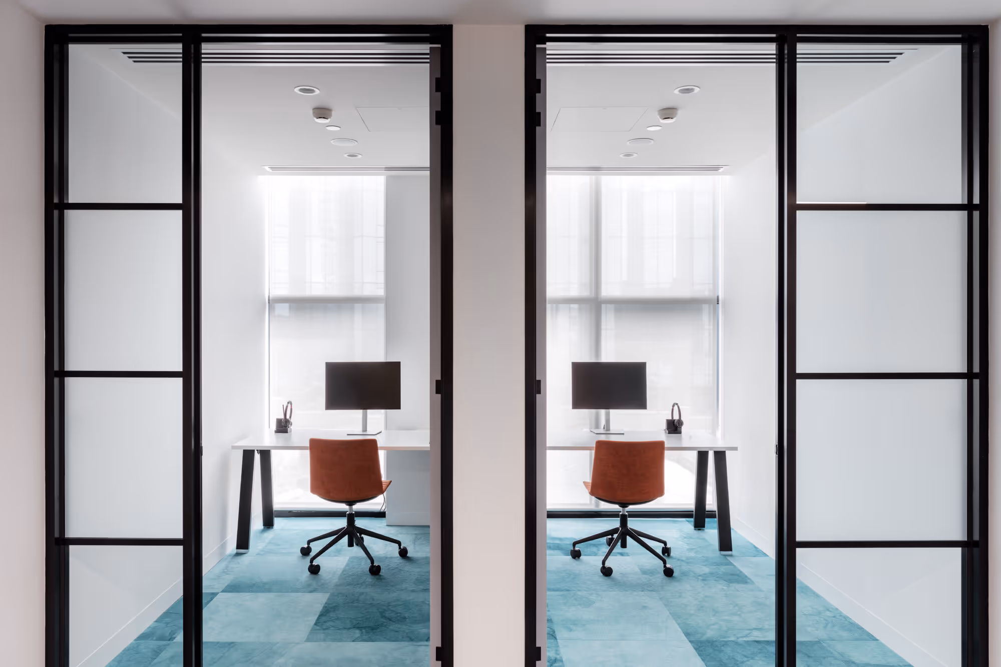 Two minimalist office cubicles with orange chairs, black monitors, and blue carpet behind glass doors.