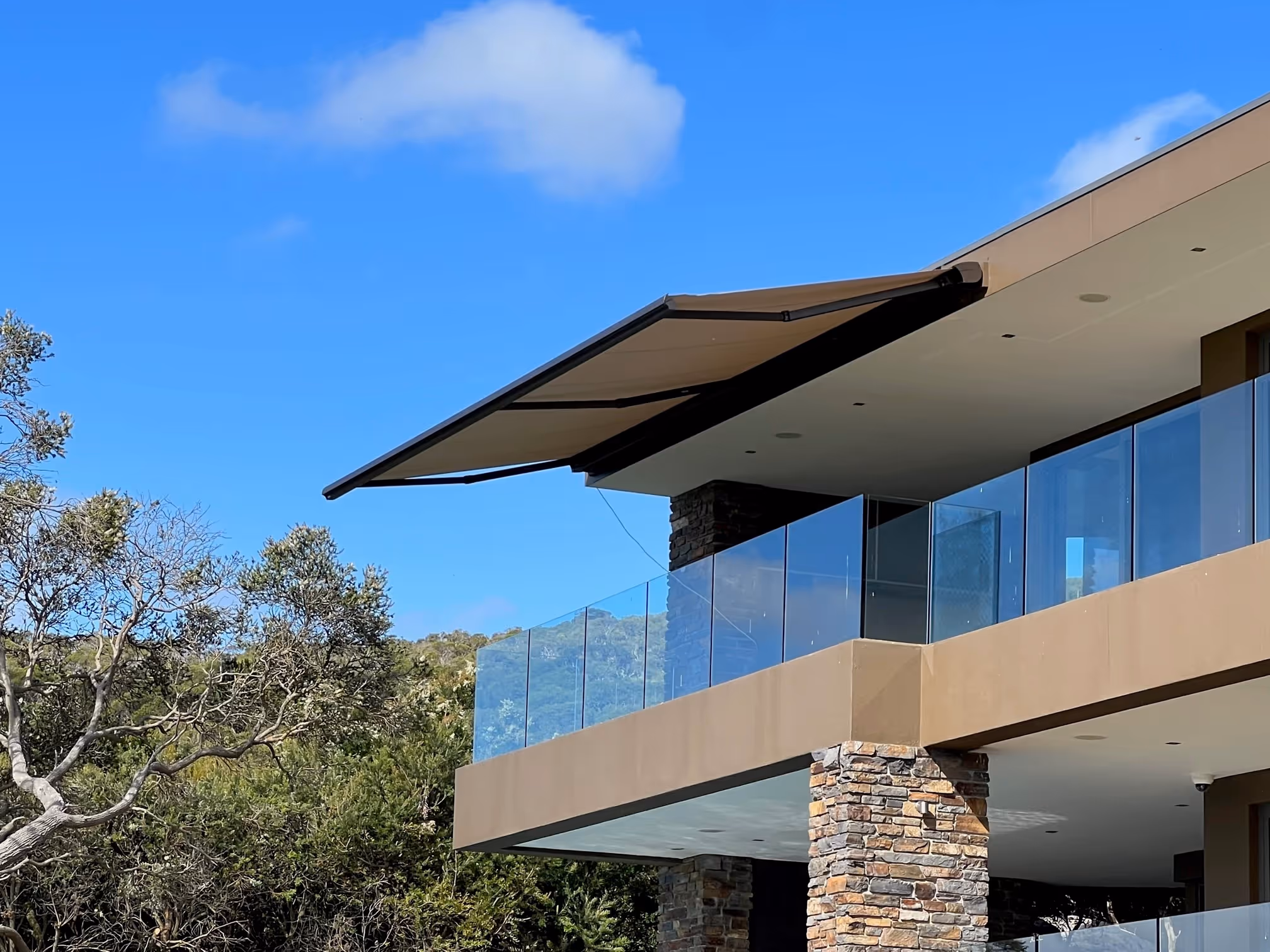 Modern house balcony with glass railing, stone pillars, and extended beige retractable awning under blue sky.