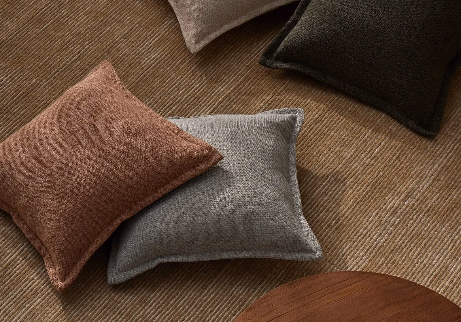 Four textured square pillows in brown, gray, dark green, and beige on a ribbed brown rug.
