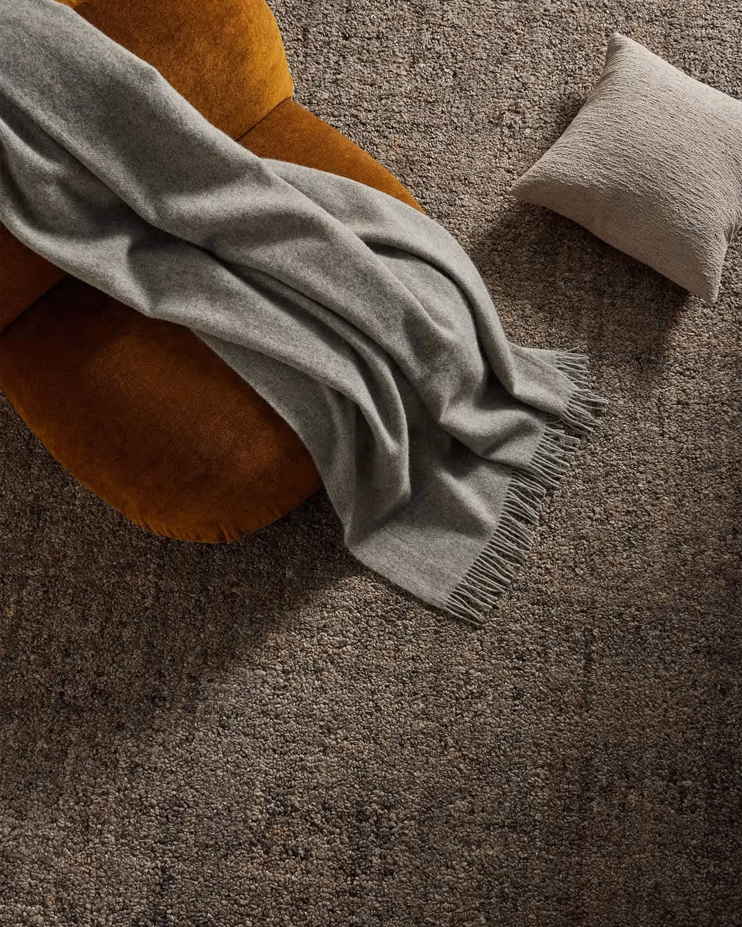 Gray fringed blanket draped over a brown cushion on a textured brown carpet with a small pillow.