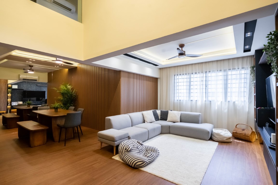 Open-plan living and dining room with warm wood floors, grey sectional sofa, and a full-length fluted wood panel feature wall behind the dining area, lit by cove lighting and sheer-curtained windows.