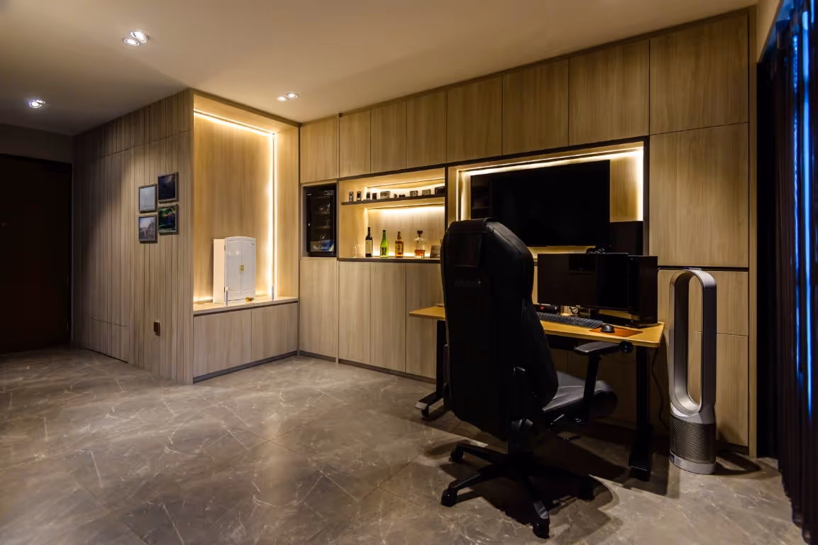 Modern home office with wall-to-wall wood cabinets and lit display niches, desk with dual monitors and gaming chair, tower fan, and polished black marble floor with subtle veining.