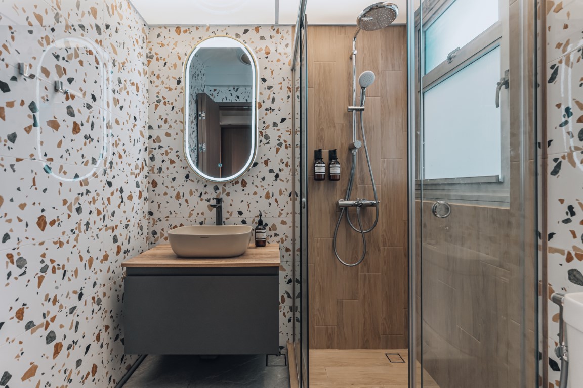 Small modern bathroom with terrazzo wall tiles, an oval backlit mirror, a beige vessel sink on a wood vanity, and a glass shower with wood-look tiles and a rain shower.