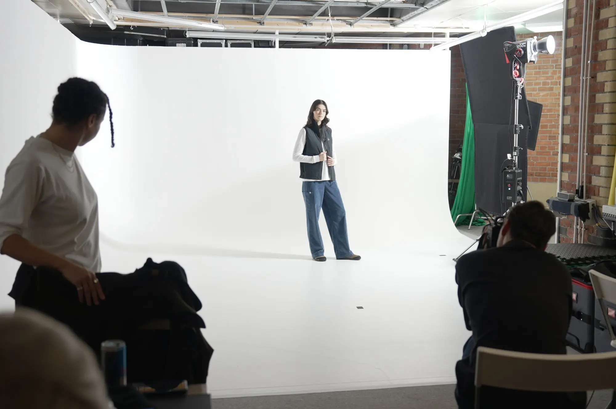 Model posing in a black vest and blue pants in a photography studio with white backdrop while a photographer takes pictures and another person watches.