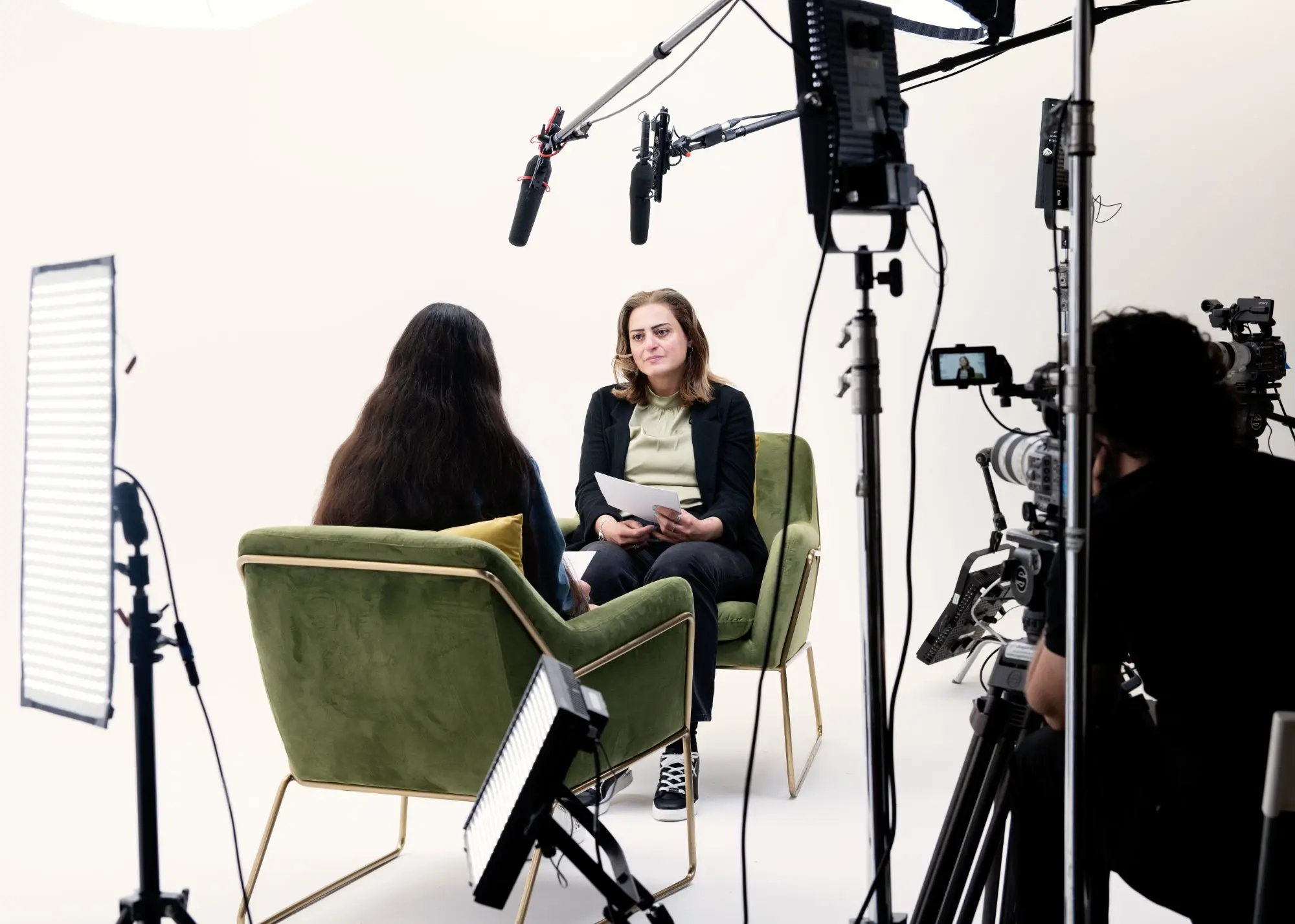 Two women seated facing each other in green chairs having an interview in a studio setup with professional lighting, microphones, and cameras.