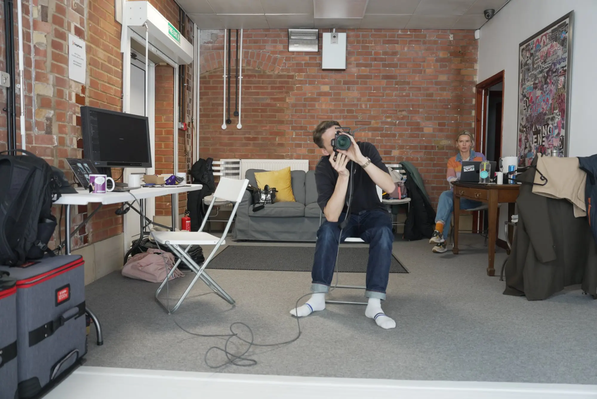 Man sitting on a chair taking a photo with a camera in a room with exposed brick walls and a gray couch in the background, while a woman sits at a wooden table with a laptop.