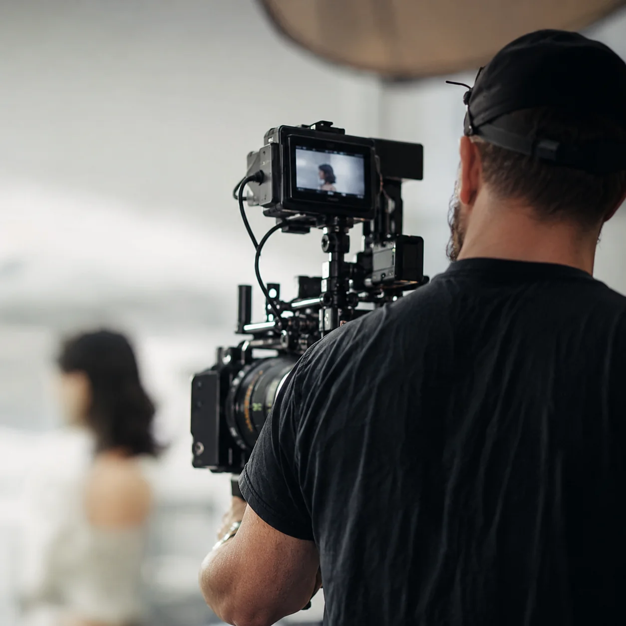 Camera operator in a black cap and shirt filming a woman with dark curly hair, viewed from behind with the camera's monitor displaying her framed shot in the foreground.