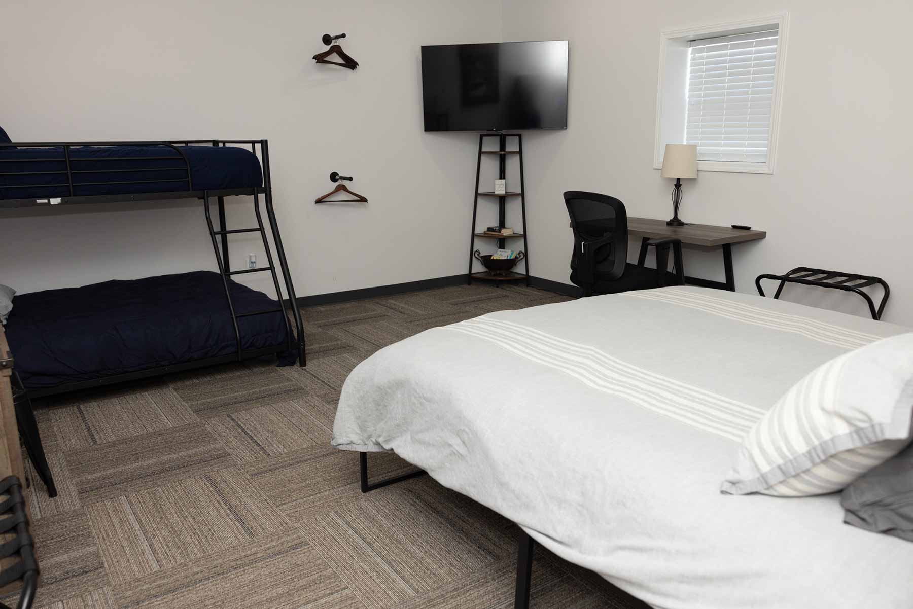 Room with a bunk bed with navy blue bedding, a queen bed with light gray bedding, a desk with a black chair and lamp, a wall-mounted flat-screen TV, and two wooden hangers on the wall.