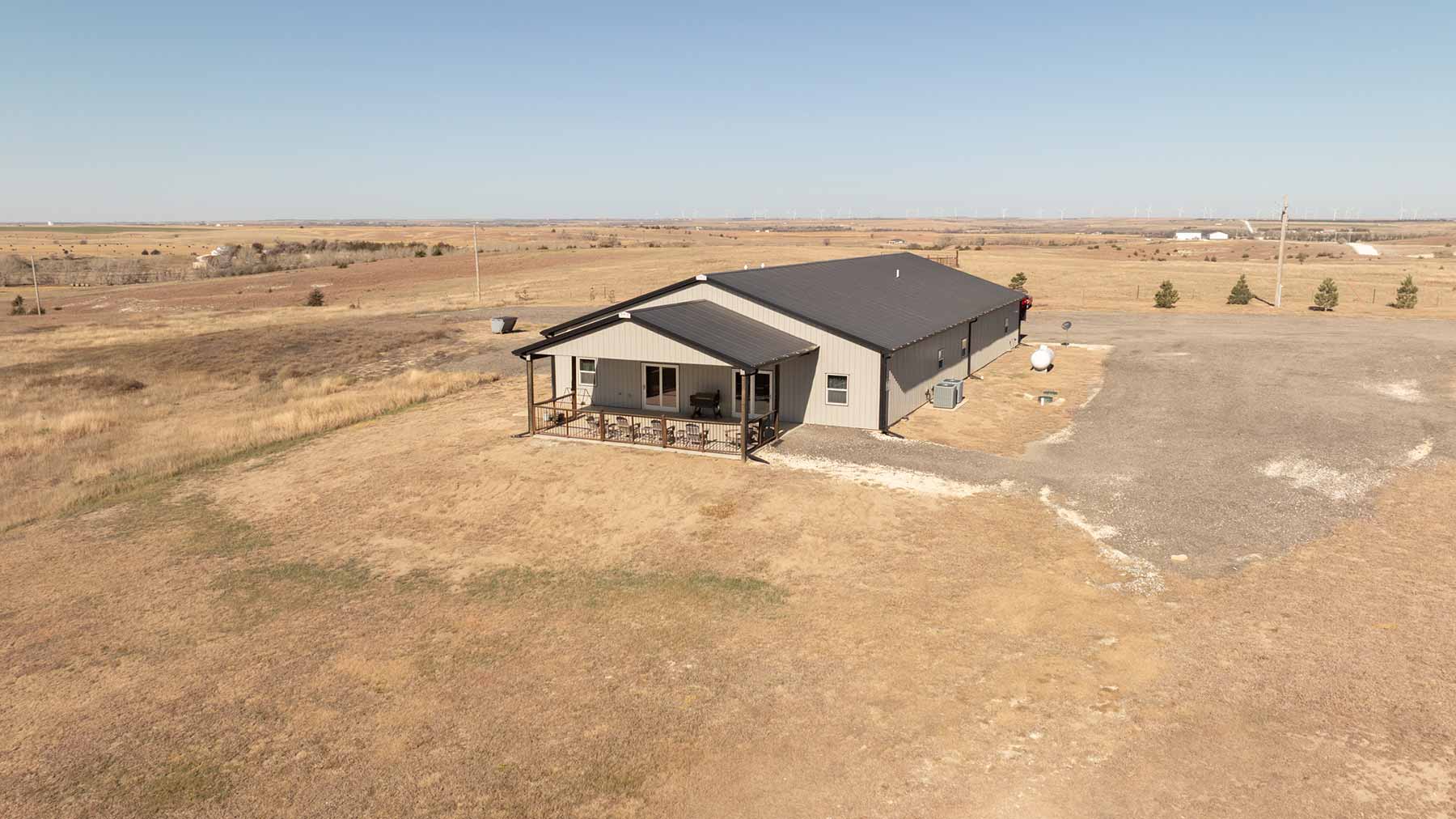 Single-story beige metal building with a covered porch and chairs, situated in a dry rural landscape under clear sky.