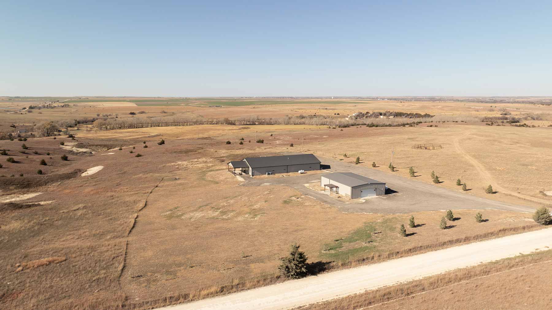 Two large rectangular buildings on a dry grassland with sparse trees and a dirt road in the foreground under a clear blue sky.